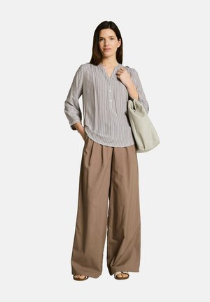 Frau trägt eine leichte gestreifte Bluse und weite beige Hosen, hält eine große weiche beige Tasche über der Schulter und steht ruhig.