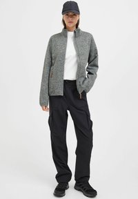 Männergrauer Fleecepullover mit hohem Kragen, Reißverschlüssen und Seitentaschen, kombiniert mit schwarzen Cargo-Hosen und dunklen Sneakers.