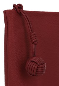 Geantă roșie din piele cu suprafață texturată, prevăzută cu închidere cu șnur legat în nod și detaliu rotund cu ciucure.