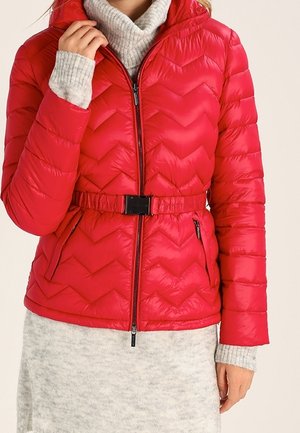 Femme portant une veste matelassée rouge avec une taille ceinturée, une fermeture éclair et des poches zippées, par-dessus une robe pull légère gris clair.