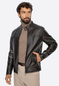 Mężczyzna z brodą, ubrany w czarną skórzaną kurtkę, beżowy golf i spodnie, z jedną ręką w kieszeni kurtki, patrzący w bok.