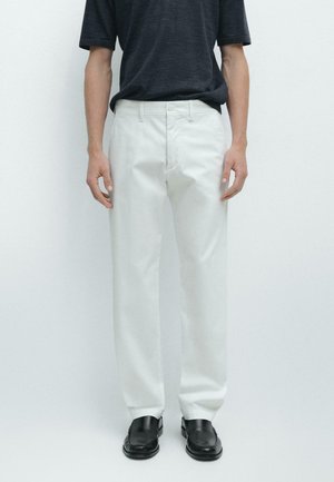 Persona che indossa pantaloni bianchi a gamba dritta, mocassini neri e una camicia scura a maniche corte, in piedi contro uno sfondo chiaro e uniforme.