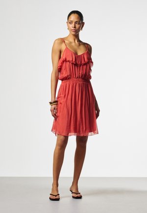 Frau steht und trägt ein ärmelloses korallenfarbenes Kleid mit Rüschen und elastischer Taille, kombiniert mit Armbändern und schwarzen Sandalen, vor neutralem Hintergrund.