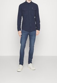 Granatowa koszula z długim rękawem, z dwiema kieszeniami na piersi, z zapięciem na guziki, w połączeniu z dopasowanymi niebieskimi dżinsami i białymi trampkami. Prosty, codzienny strój.