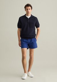Polo navy a maniche corte con tessuto a maglia texturizzato e taschino, abbinato a pantaloni corti a righe blu. Sneakers bianche completano l'outfit.