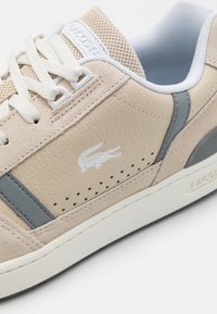 Basket Lacoste beige et grise avec lacets blancs, panneau latéral perforé et logo crocodile sur cuir texturé.