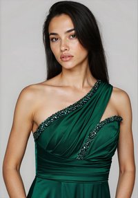 Giovane donna con lunghi capelli scuri che indossa un abito di raso verde smeraldo con dettagli di perline su una spalla, su uno sfondo semplice.