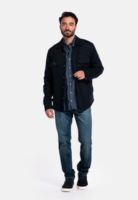 Man in een donkerblauwe jas over een blauwe blouse, gekleed in blauwe jeans en zwarte sneakers, staand tegen een effen lichte achtergrond.