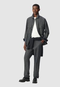 Giacca di lana grigia a trama con doppie tasche sul petto, abbinata a pantaloni coordinati e scarpe in pelle nera. Maglione di lana blu navy drappato in vita.