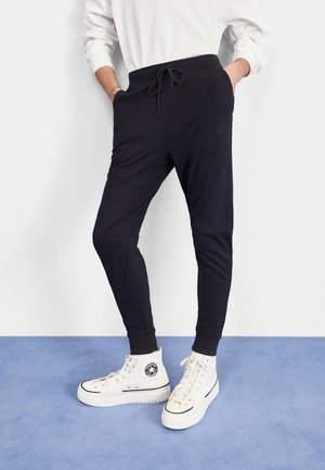 Person, die schwarze, schmal zulaufende Sweathosen mit Kordelzug und weiße High-Top-Sneakers trägt, steht auf blauem Boden.