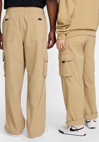 Beżowe spodnie typu cargo z dużymi bocznymi i tylnymi kieszeniami, elastycznym paskiem w talii i luźnym krojem; noszone z białymi trampkami z czarnymi elementami.