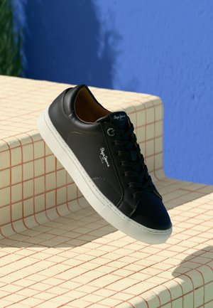 Black Pepe Jeans sneaker with white sole, displayed on beige tiled steps with a blue wall background.