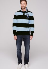 Pullover a righe blu chiaro e nero con colletto, dotato di un logo e testo. Indossato con jeans scuri e sneakers bianche.
