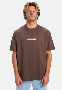 Brun bomulds T-shirt med et rektangulært rødt Quiksilver-logo på brystet. Korte ærmer, afslappet pasform og rund hals.
