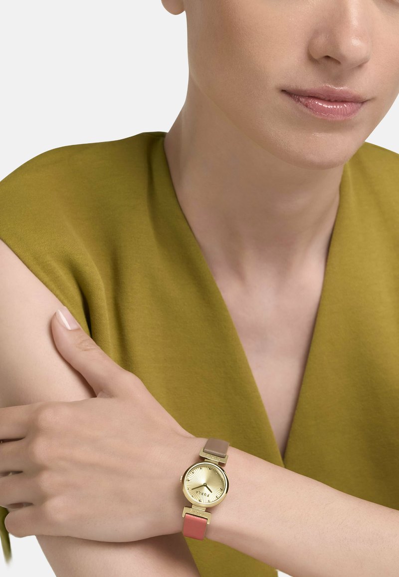 Gros plan sur une personne portant une montre en or avec un bracelet en cuir bicolore, vêtue d'un haut sans manches vert olive.