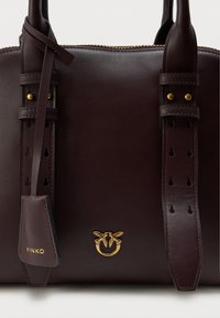 Καφέ δερμάτινη τσάντα χειρός με διπλές λαβές, χρυσά μεταλλικά στοιχεία και διακοσμητικές ζώνες με κοψίματα. Έντονη λεπτομέρεια λογότυπου στο μπροστινό μέρος.