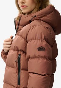 Chaqueta de plumón marrón con capucha, con un bolsillo con cremallera en la parte frontal, puños elásticos y un parche con el logo en la manga. Textura suave y brillante.