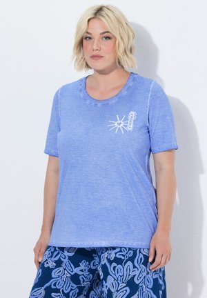 Lyshåret kvinde iført en lyseblå T-shirt med sol- og hjerte-design og mørkeblå mønstrede bukser, stående mod en hvid baggrund.