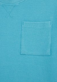 Svetlo modra pletena majica z teksturiranim tkanjem in majhno všitno žepno na levi strani. Okrogel izrez.
