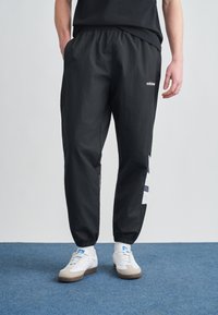 Černé sportovní kalhoty s elastickým pasem, bočními kapsami a kontrastními pruhy po stranách. Vyrobené z lehkého, hladkého materiálu.
