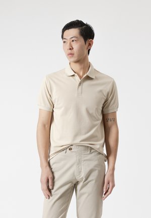 Joven de pie, vistiendo una camisa polo beige y pantalones a juego, mirando hacia un lado sobre un fondo blanco liso.