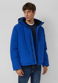 Niebieska pikowana kurtka puchowa z zamkiem z przodu, kapturem i kieszeniami. Wyróżnia się pikowanym wzorem oraz miękką, teksturowaną tkaniną.