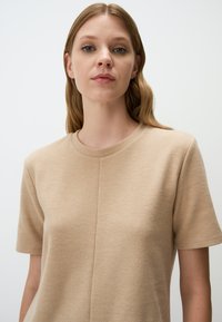 Beige kurzärmliges Stricktop mit Rundhalsausschnitt, das eine dezente strukturierte Oberfläche und Nahtdetails auf der Vorderseite aufweist.