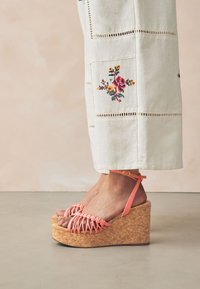 Koralfarvede kilehøje sandaler med vævede stropper, korksål, ankelrem med spænde, kombineret med lyse broderede bukser.