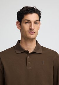 Brun polo skjorta med en texturerad tyg, kort krage och tre knappar, med en liten logotyp broderad på vänster bröst.