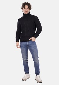 Maglione a collo alto in maglia nera con una consistenza spessa, abbinato a jeans azzurri e sneakers bianche, in piedi su uno sfondo neutro.