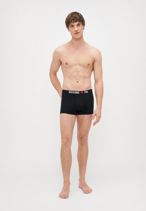 Fekete boxer alsónadrág puha derékpánttal, rajta fehér és piros "MOSCHINO ❤️ YOU" felirattal. Az anyaga sima, szorosan illeszkedik. A modell mezítláb áll.