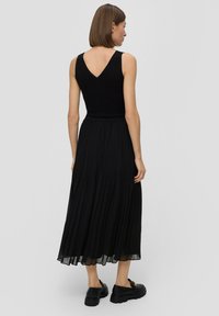 Zwarte geplooide maxi-jurk met een aansluitend lijfje en V-achterkant. Heeft een vloeiende rok en een ronde zoom. Gepaard met zwarte schoenen.