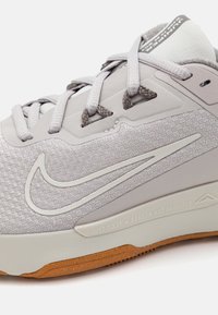 Zapatilla deportiva gris claro con parte superior texturizada, cordones en un gris más oscuro y suela de goma. Presenta un logotipo prominente y texto grabado.