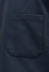 Σκούρο μπλε ύφασμα με ραμμένη ορθογώνια τσέπη και ορατό ραφάκι φερμουάρ στην αριστερή πλευρά.