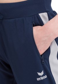 Marineblauwe sportbroeken met een ritszak aan de zijkant, elastische tailleband en een contrasterende grijze streep. Bevat een logo op het onderbeen.