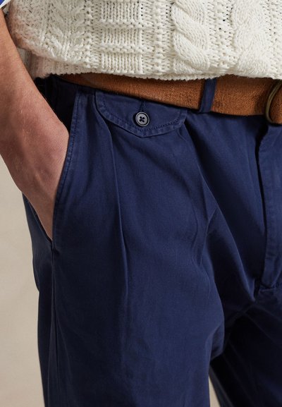 Ruka u džepu mornarskoplavih hlača s džepom na preklop s gumbom, smeđi pojas i bijeli pulover pleten uzorak kabela vidljiv u području struka.
