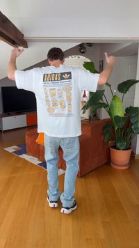 Personne portant un t-shirt blanc Adidas avec un imprimé graphique, levant les deux bras dans un salon avec des plantes et un canapé orange.