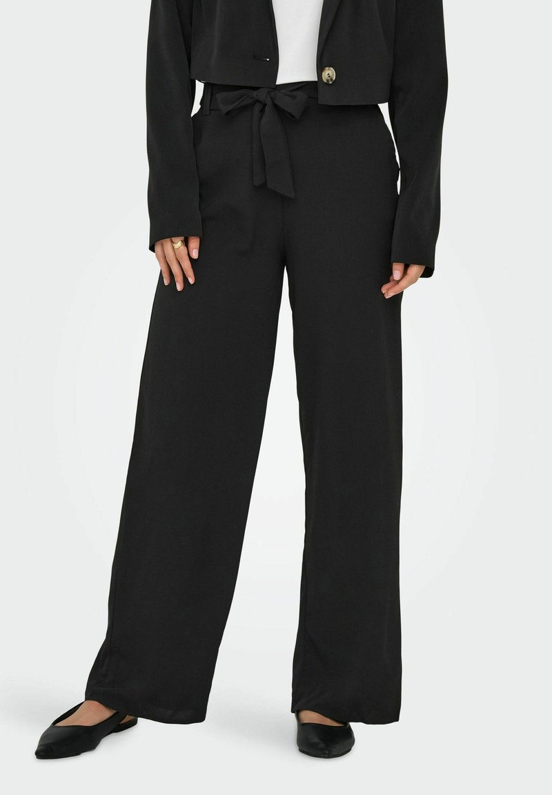 Pantalon noir à jambes larges avec une texture lisse, doté d'une taille nouée et d'une veste courte. Les chaussures sont des ballerines noires simples.