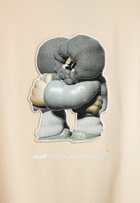 Tela beige con un diseño bordado detallado de un personaje con elementos grises, extremidades esponjosas y una apariencia texturizada. "MWM x mod wave movement" está impreso debajo.