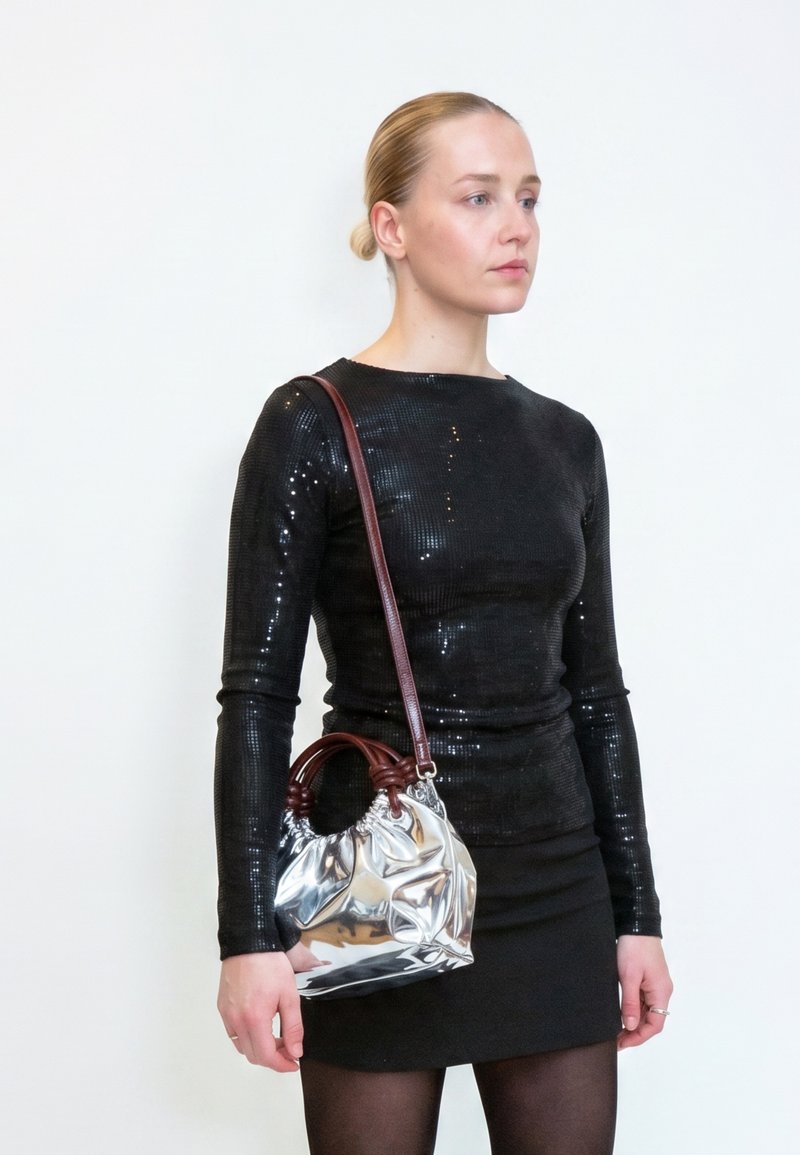 Femme aux cheveux en chignon porte une robe noire à manches longues pailletée et tient un sac à main argenté brillant avec des poignées et une bandoulière marron.