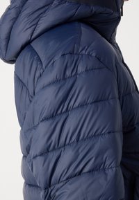 Veste matelassée bleu marine avec capuche, gros plan sur l'épaule et la partie supérieure du bras.