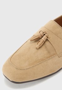 Beige Wildleder Slipper mit runder Spitze, dekorativen Knoten an der Vorderseite und kontrastierenden Nähten entlang der Ränder.