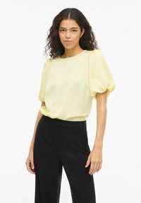 Blouse jaune à manches courtes bouffantes, tissu lisse, encolure ronde, associée à un pantalon noir, design minimaliste, sans motifs ni accents.