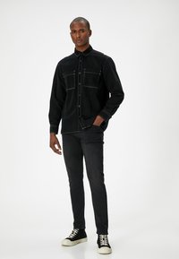 Camisa de mezclilla negra con botones y dos bolsillos en el pecho, costuras blancas en contraste y un corte relajado. Se combina con jeans negros y zapatillas negras.