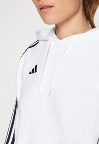 White hoodie made of soft material, features black three-stripe accents on sleeves and a small black logo on the chest.