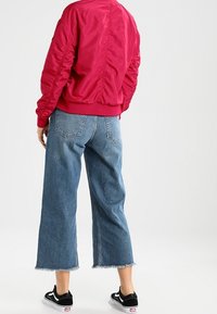 Czerwona satynowa kurtka bomberka z teksturowanymi rękawami, zestawiona z jasnoniebieskimi jeansami o kroju cropped z postrzępionymi brzegami, noszona z czarnymi trampkami.