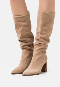 Bottes en daim taupe montant jusqu'au genou avec un design affaissé, un bout pointu et un talon en bois empilé. La texture semble douce et lisse.