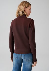 Brauner Pullover mit geripptem Kragen und langen Ärmeln, kombiniert mit hellblauen Jeans. Glatte Textur, figurbetontes Design mit einem leichten Stretch.