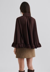 Cape marron à volants avec un col montant ; son tissu léger tombe en une ourlet évasé. En dessous, une jupe texturée marron complète la cape.