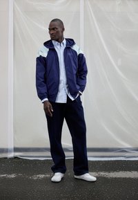 Navy windbreaker with light blue accents, zip pockets, and hood, worn over a light blue shirt and dark denim pants, complemented by white sneakers.